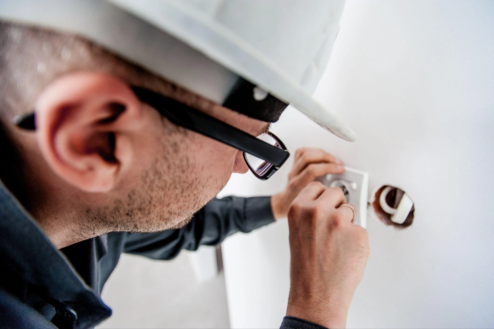 Technician in a white hard hat and glasses fixing a wall socket during a home repair visit